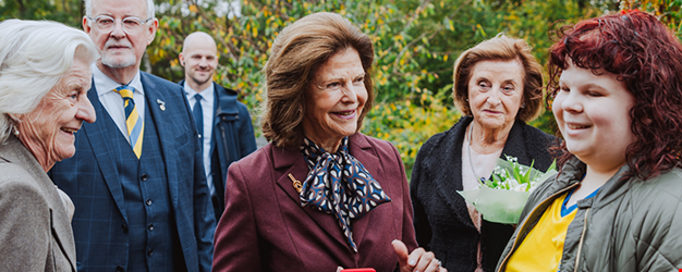 En grupp människor i samtal utomhus. I mitten står en äldre kvinna i vinröd kavaj och scarf som ler och pratar med en ung person i gul tröja. Runt omkring står flera andra vuxna, varav en håller en bukett blommor. Träden i bakgrunden antyder att det är höst.