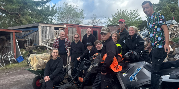En grupp personer står och sitter tillsammans utomhus framför skjul och vedtravar. 
