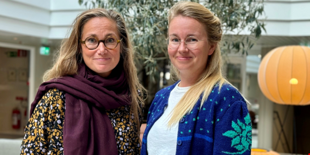 Två kvinnor står tillsammans inomhus i ett ljust, modernt rum med gröna växter i bakgrunden. Den ena bär glasögon, en mörk lila halsduk och en mönstrad blus i gult och svart. Den andra har långt blont hår, glasögon och en blå kofta med grön mönsterdetalj över en vit tröja. Båda ser in i kameran och ler svagt.