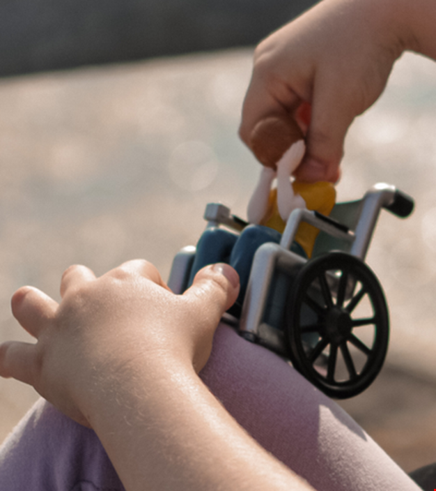 Bild på ett barns händer. Barnet leker med en leksaksgubbe som sitter i rullstol.