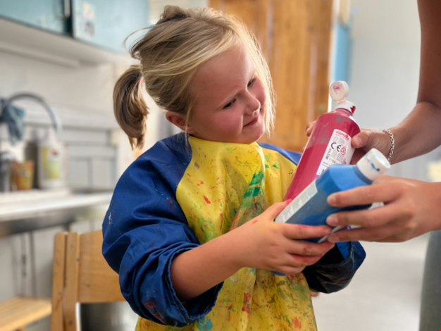 Flicka iklädd förkläde håller i en flaska med röd färg och en flaska med blå färg. 