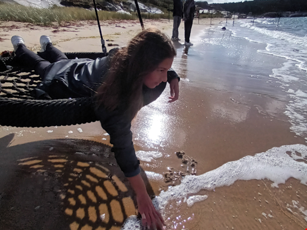 Flicka ligger på en gunga på stranden och doppar handen i havet. 