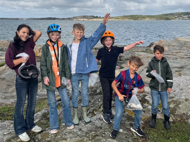 Sex barn i hjälmar står vid havet och vinkar mot kameran.