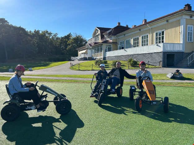 Barn och vuxna sitter i trampbilar framför Gula huset på Ågrenska.