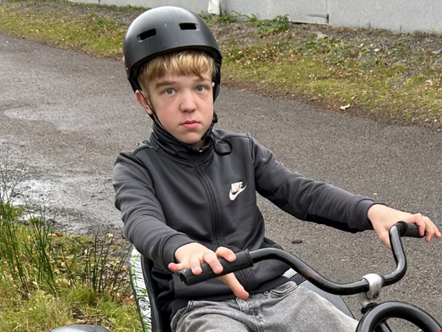 Pojke med en svart hjälm sitter på en trampbil och tittar in i kameran. 