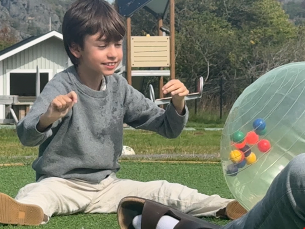 Pojke rullar en stor genomskinlig boll  sittandes mellan sig och en vuxen.