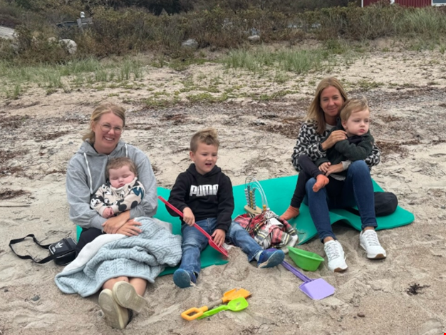 Två vuxna och tre barn sitter på en filt på sandstranden. 