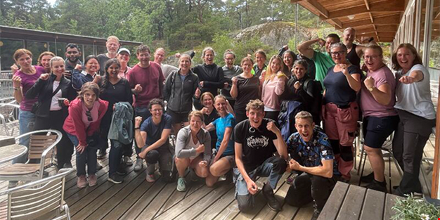 En grupp människor står samlade utomhus på en träaltan. De är glada, gör starka poser och tittar mot kameran.