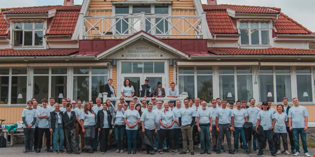 En stor grupp personer i ljusblå tröjor står samlade framför Ågrenskas gula hus och ler mot kameran.