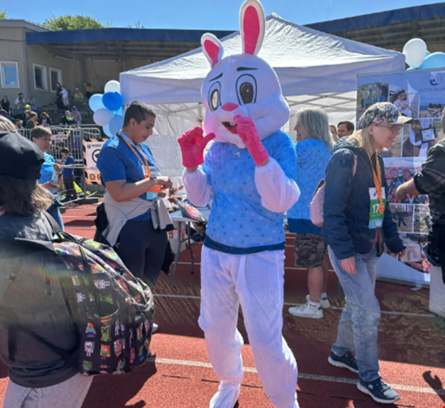 En person utklädd till en vit kanin med rosa detaljer och blå tröja står på en idrottsarena framför ett vitt tält.